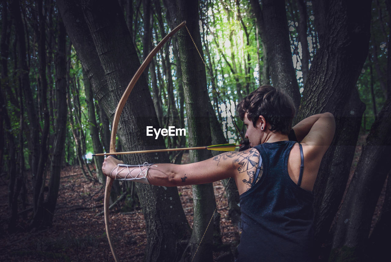 Woman holding bow and arrow while standing by tree trunk in forest