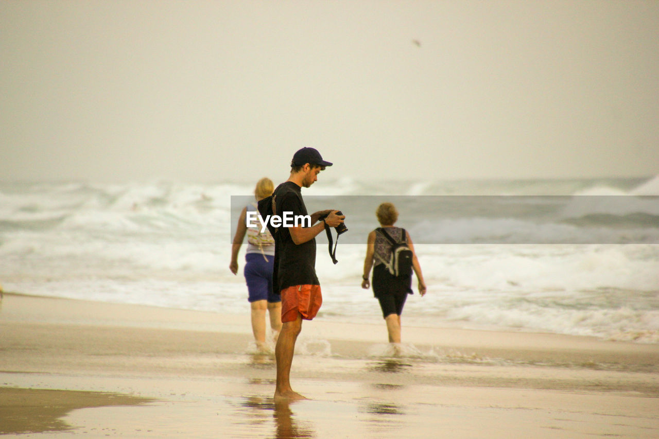 REAR VIEW OF FRIENDS ON BEACH AGAINST SKY