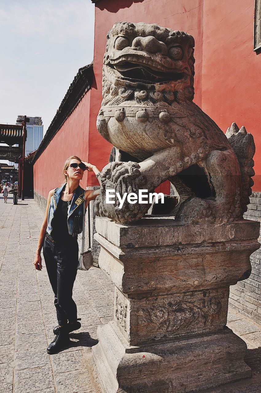 Woman standing on footpath by statue against building during sunny day