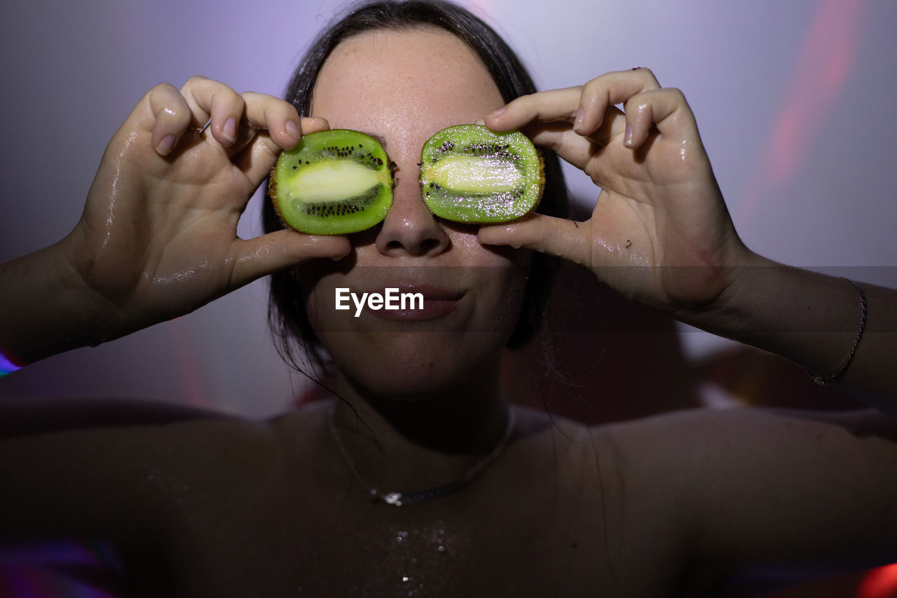 Young woman holding kiwi fruit against wall