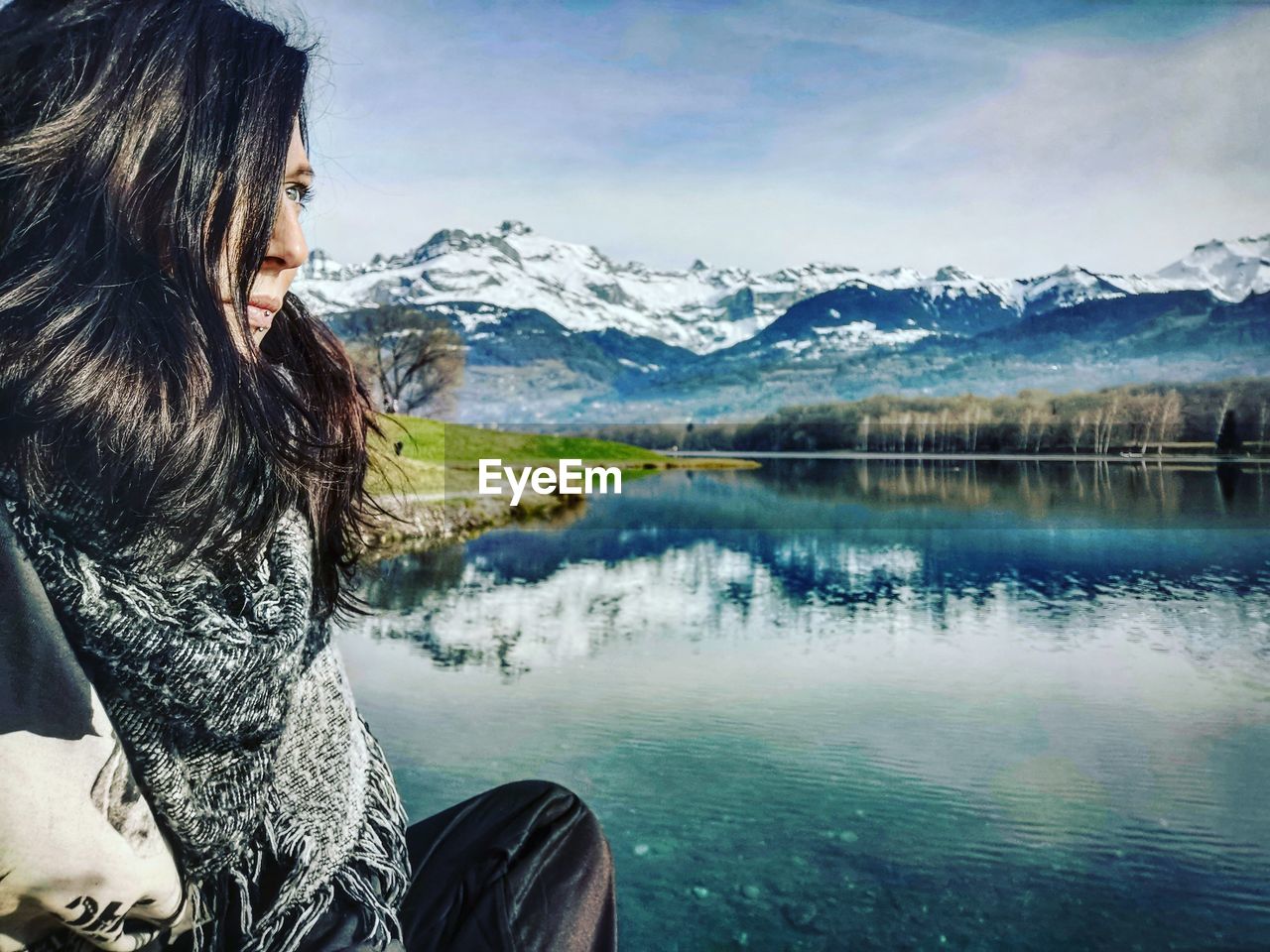 Woman looking away while sitting by lake against mountain range and sky