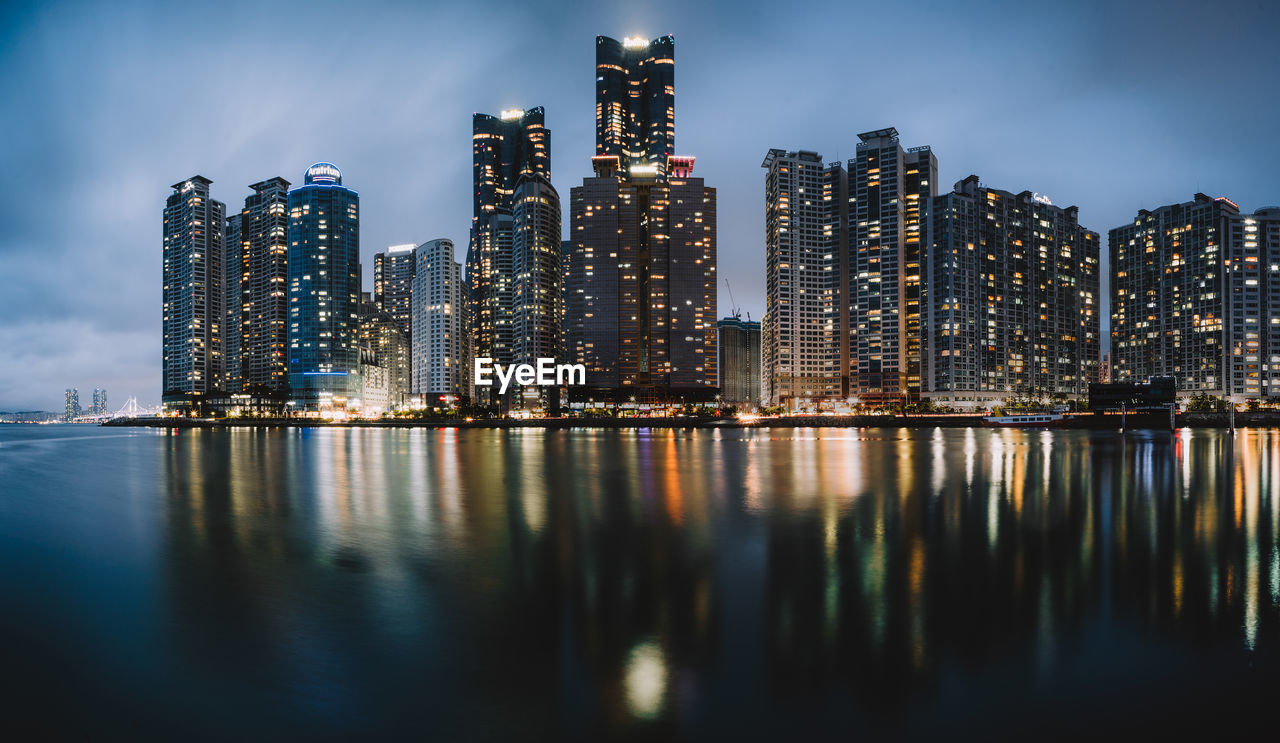 Illuminated modern buildings by river against sky at dusk