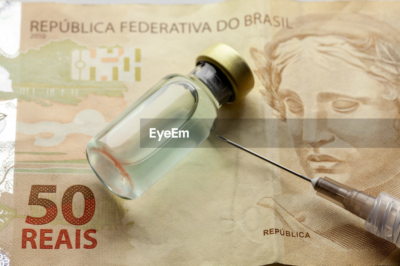 Glass vaccine bottle with liquid and syringe needle over a brazilian 50 reais bank note from close