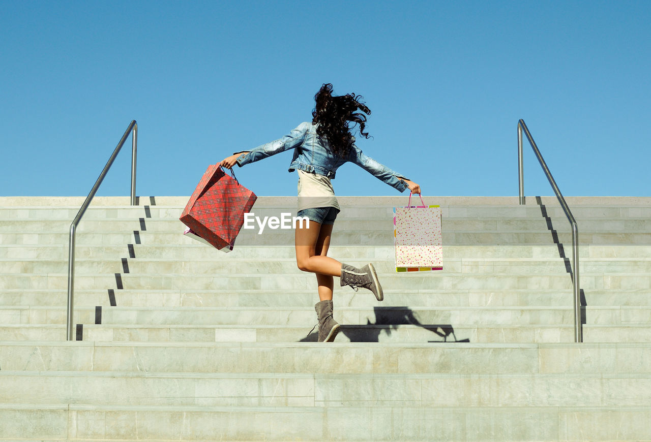 Low angle view of mid adult woman with shopping bags moving up on steps against clear blue sky