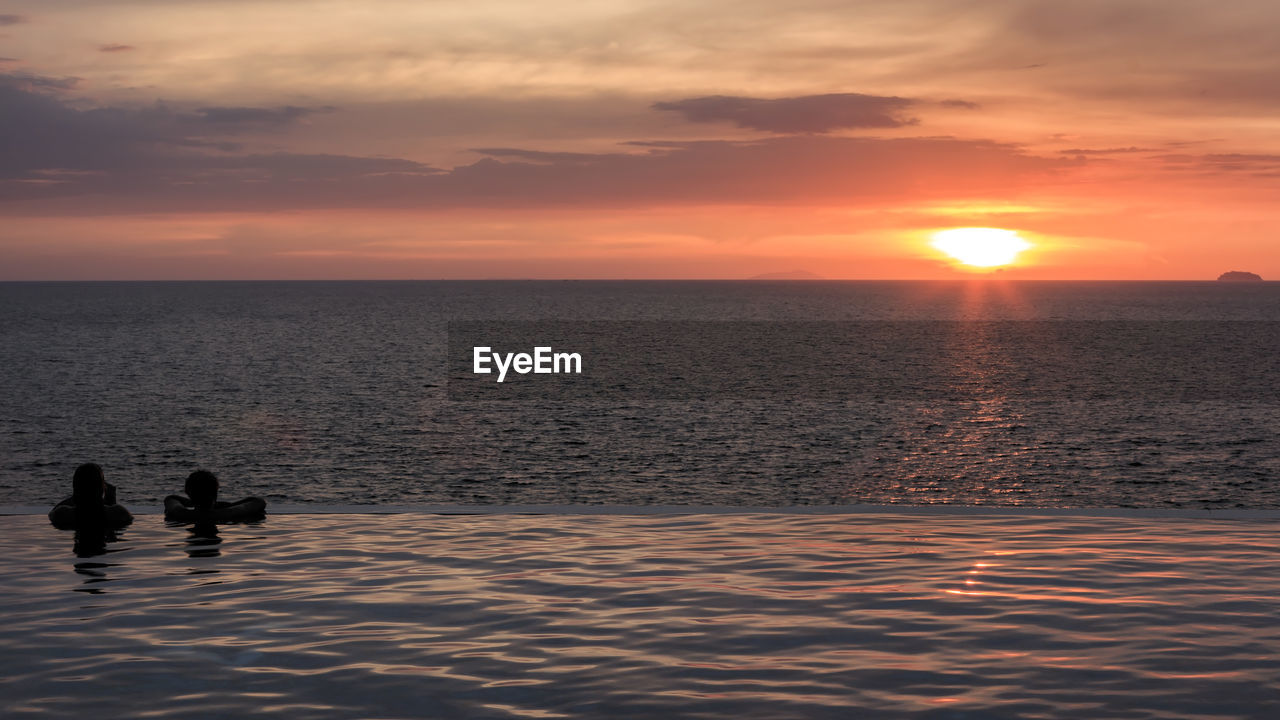 Silhouette friends in infinity pool with sea in background against orange sky