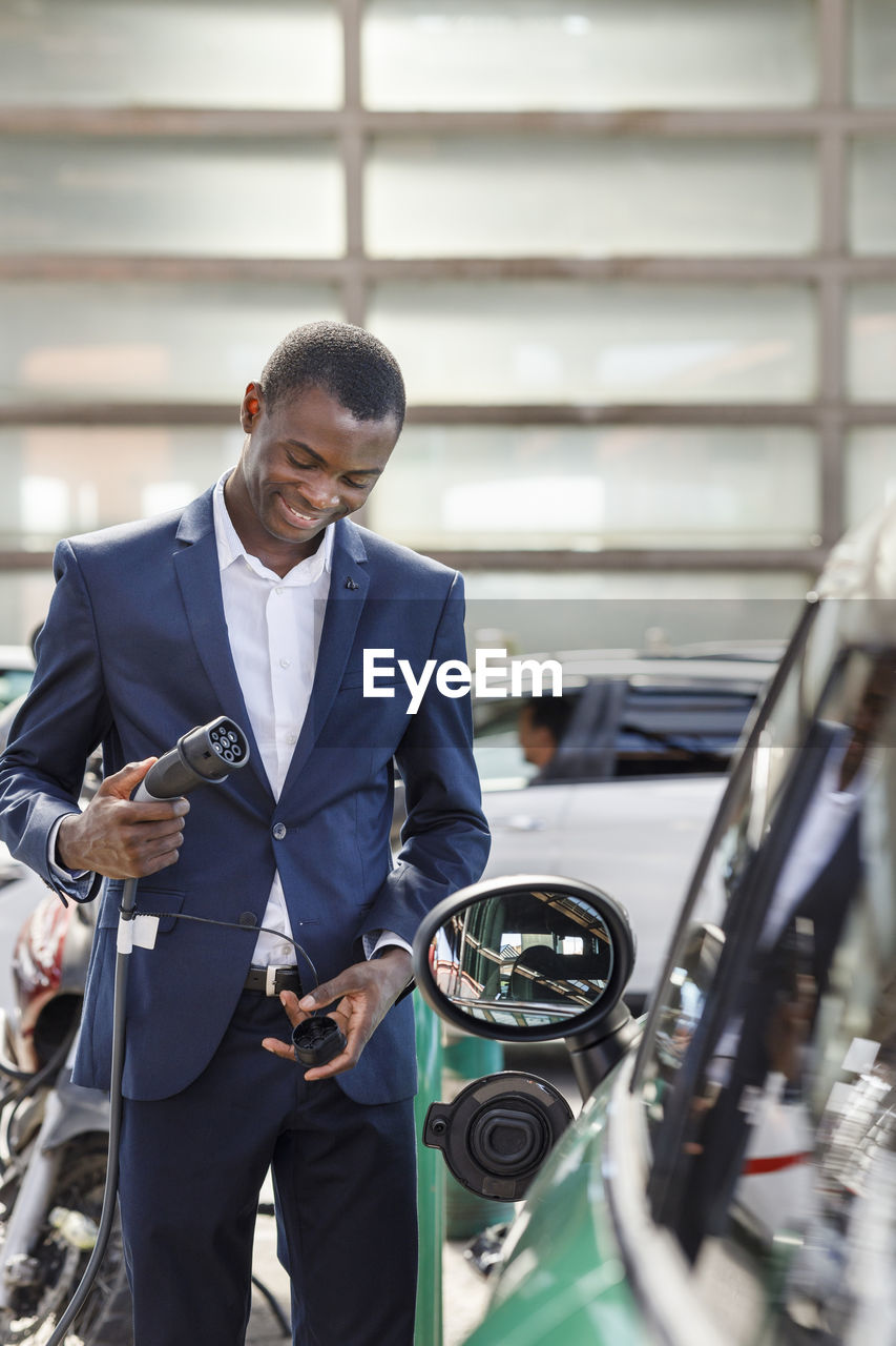 Happy businessman holding electric plug at vehicle charging station