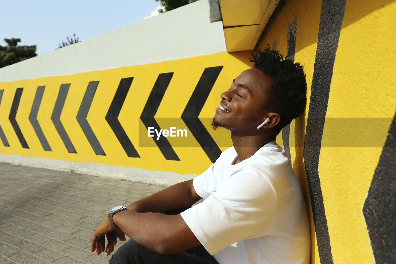 Smiling man with eyes closed listening to music sitting near wall