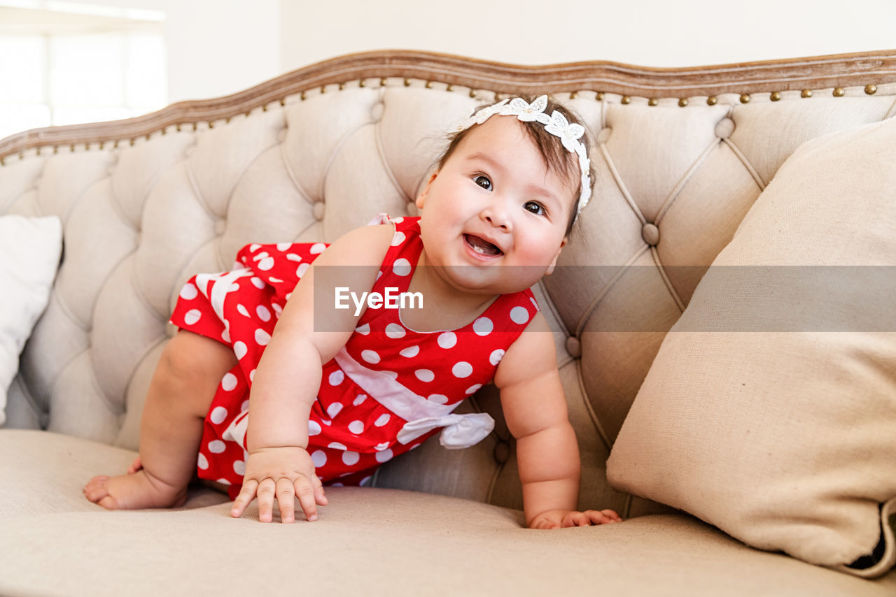 Happy asian cute baby girl in a red dress playing on the beige sofa at home