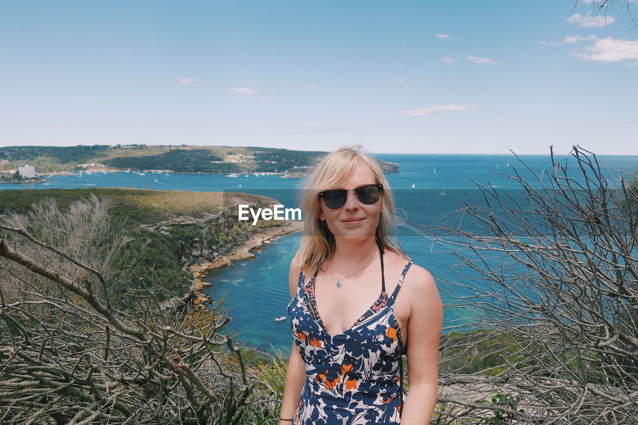 Portrait of young woman wearing sunglasses while standing against sea at beach