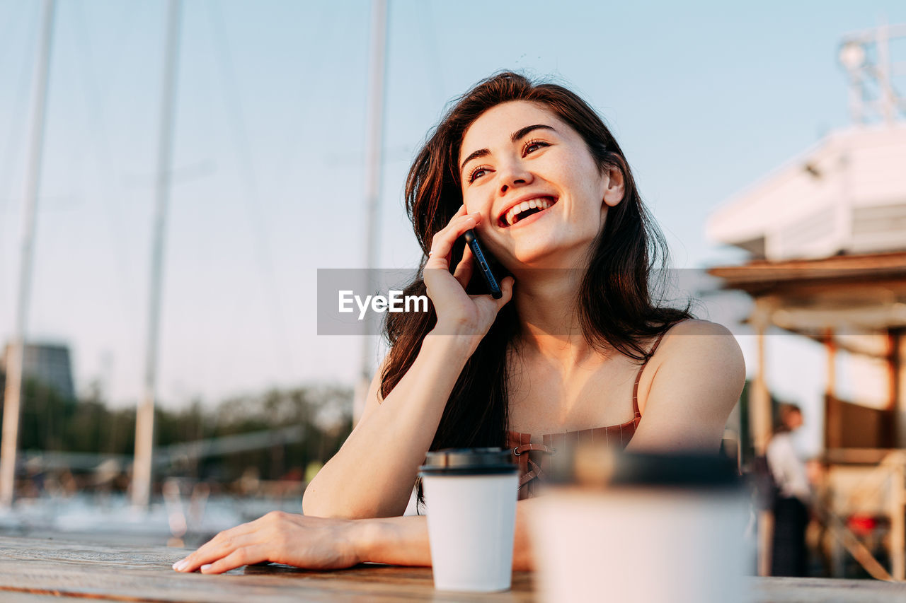 PORTRAIT OF YOUNG WOMAN USING PHONE WHILE HOLDING COFFEE CUP