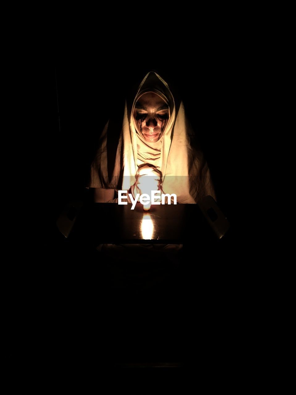 Woman with face paint sitting by illuminated light bulb in darkroom during halloween