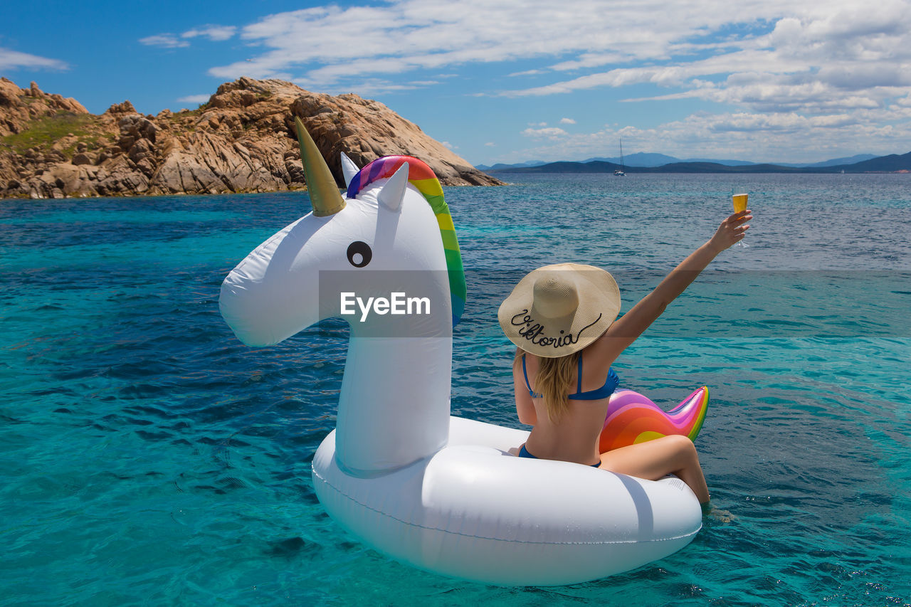Rear view of woman sitting on inflatable unicorn in aegean sea against sky