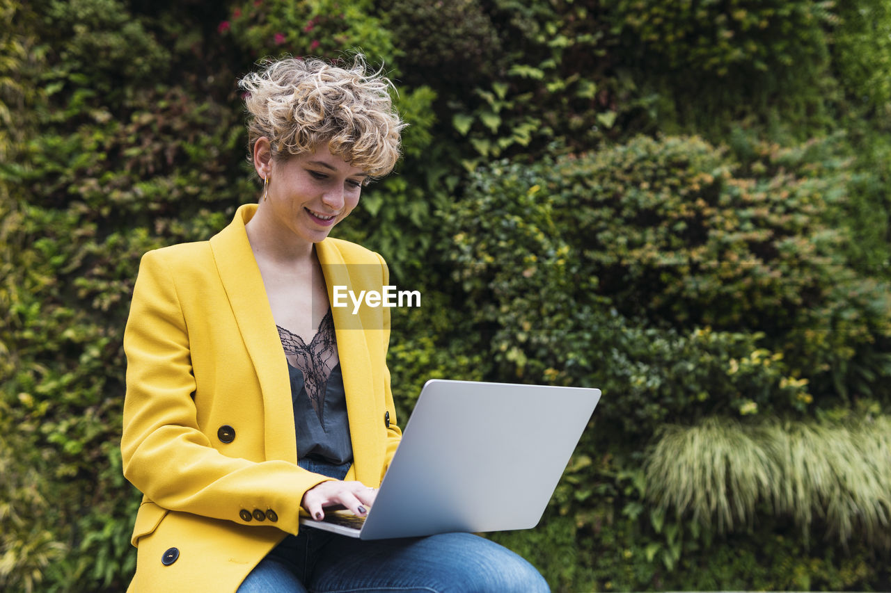 Smiling businesswoman in yellow jacket working on laptop at park