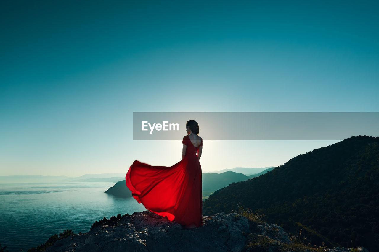 Rear view of woman looking at sea while standing on cliff against clear sky during sunset