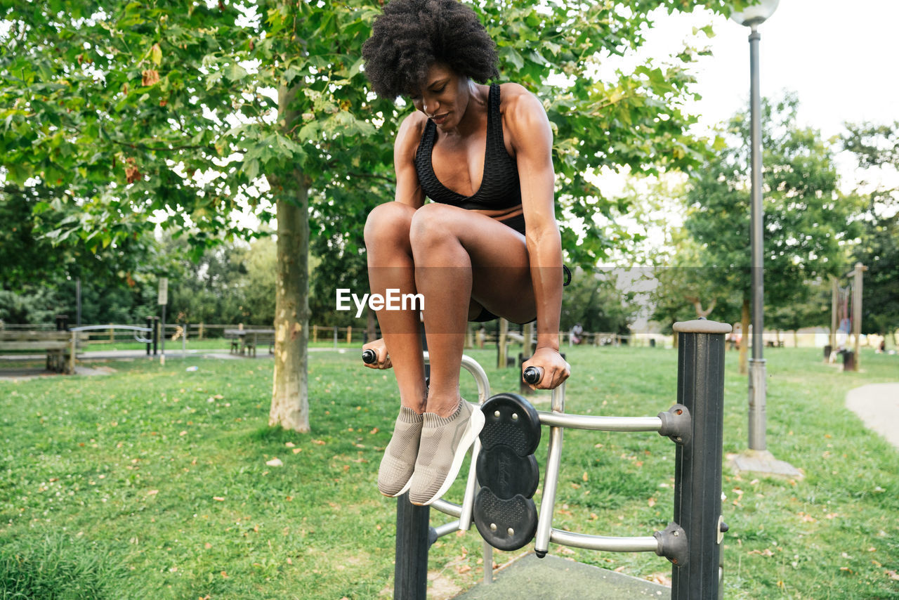Low angle of positive young curly haired black sportswoman doing push ups on parallel bars in summer park