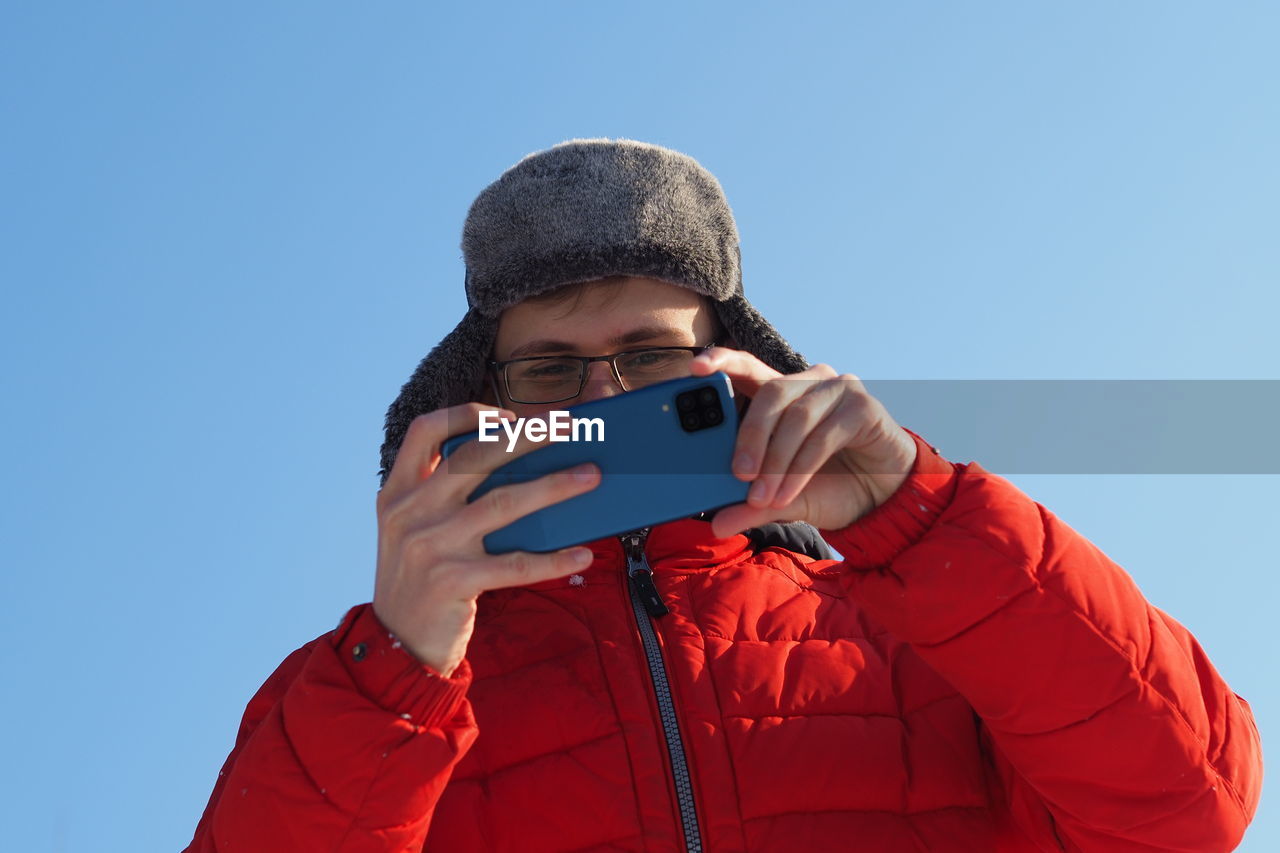 A young man of european appearance in a red jacket and a winter hat takes pictures from the phone in
