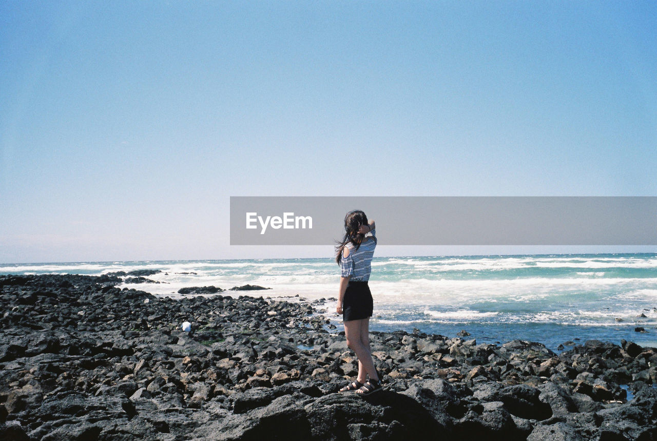 Rear view of young woman standing at seaside against clear sky