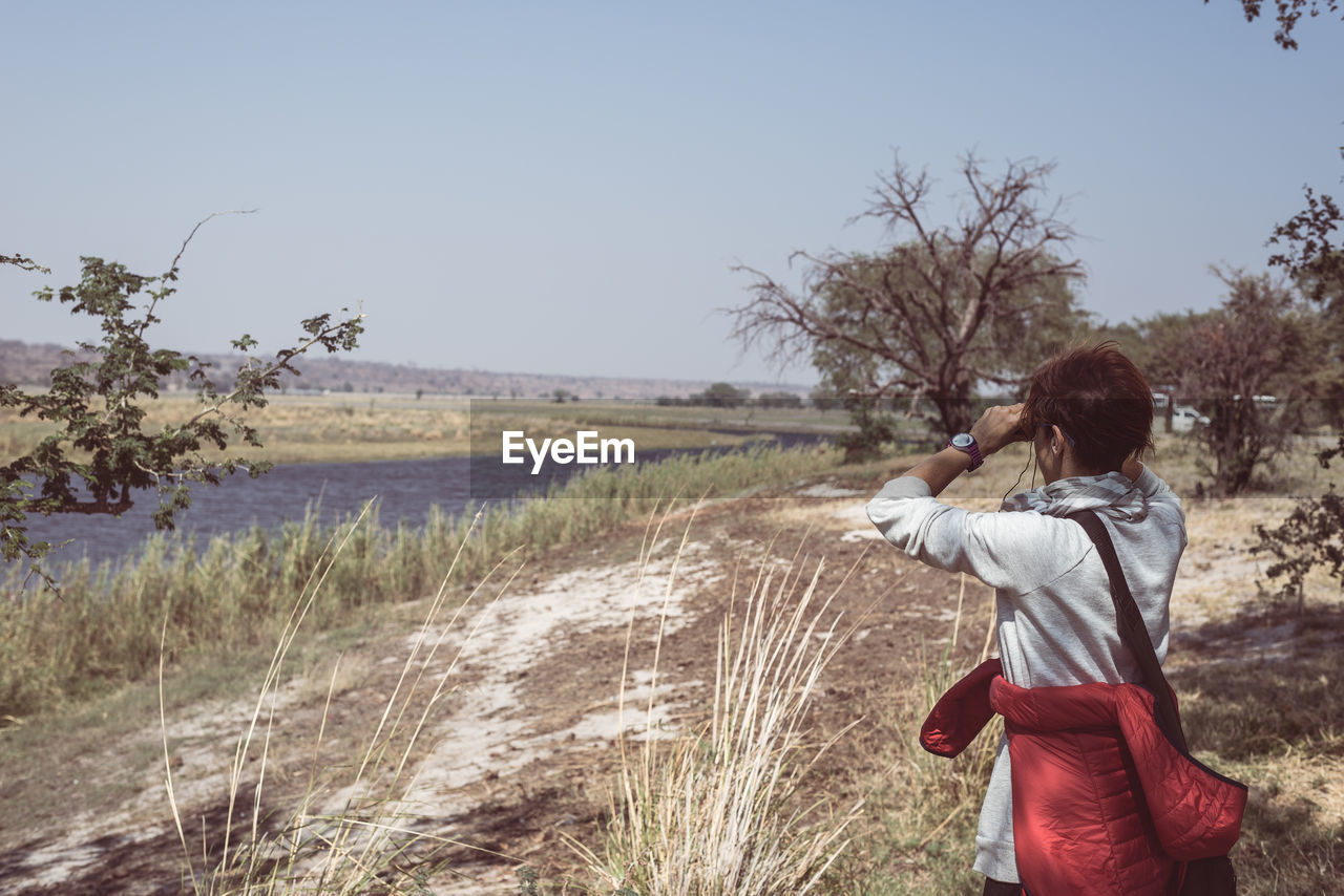 Woman looking through binoculars while standing on field against clear sky