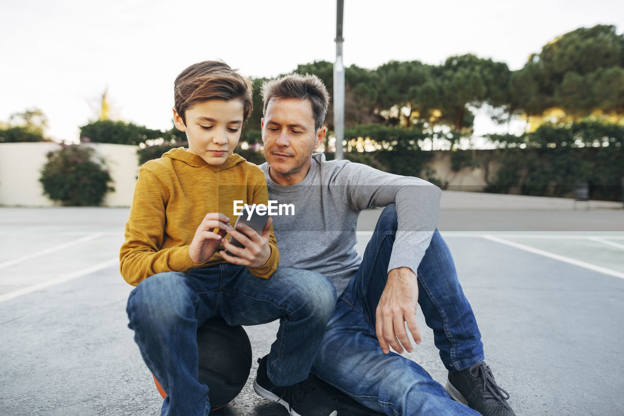 Father and son sitting on basketball outdoor court using cell phone