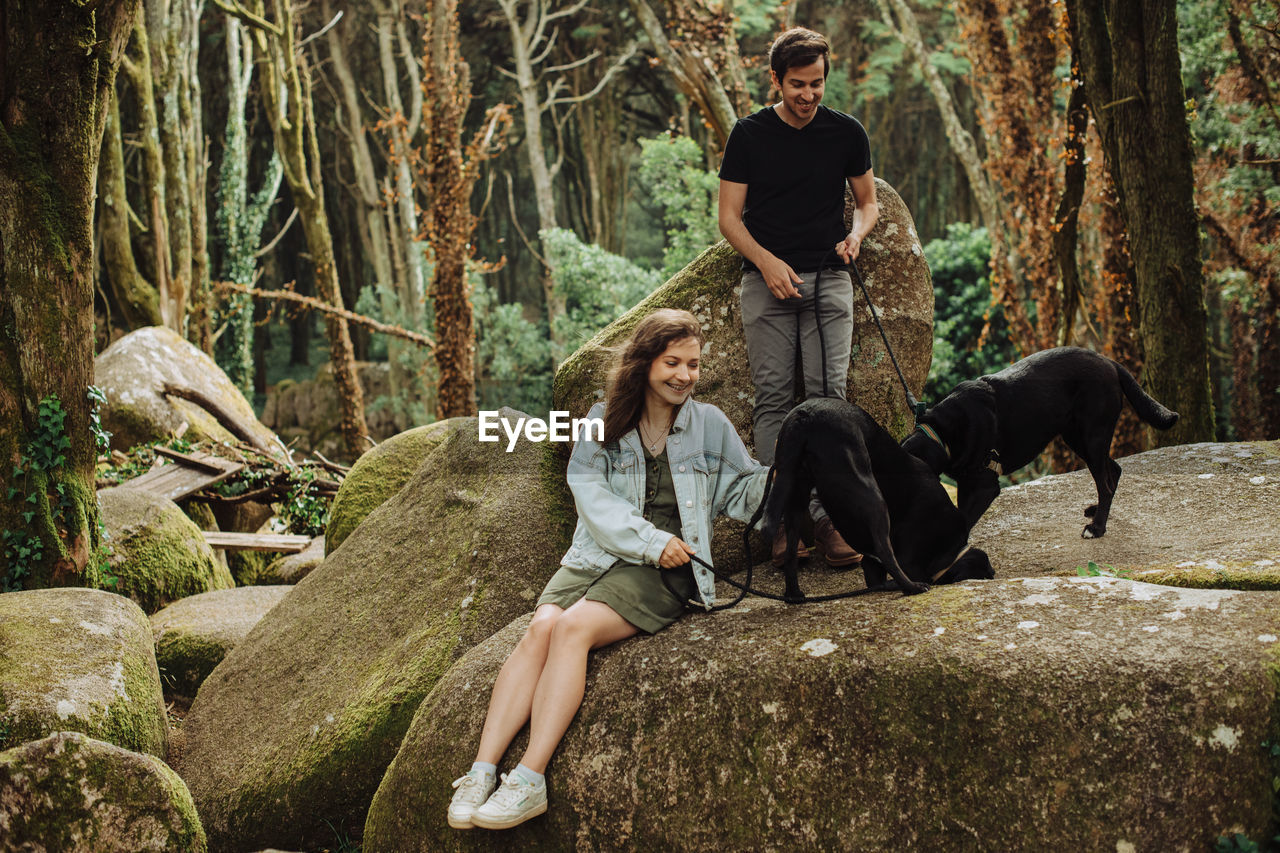 Young couple with two dogs having fun in forest while sitting on rock