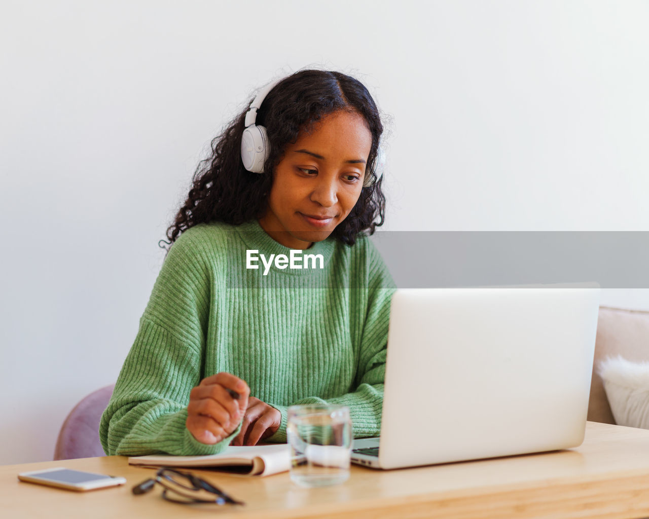 Young african-american female working on laptop in headphones while taking notes in notebook