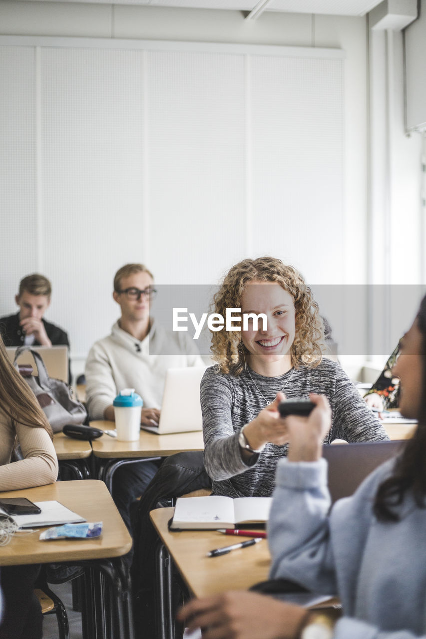 Smiling young woman giving calculator to female friend while sitting in classroom