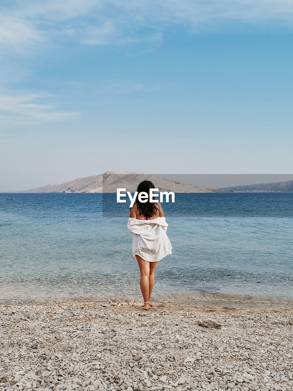 Rear view lifestyle image of stylish young woman in white shirt standing on beach and looking at sea