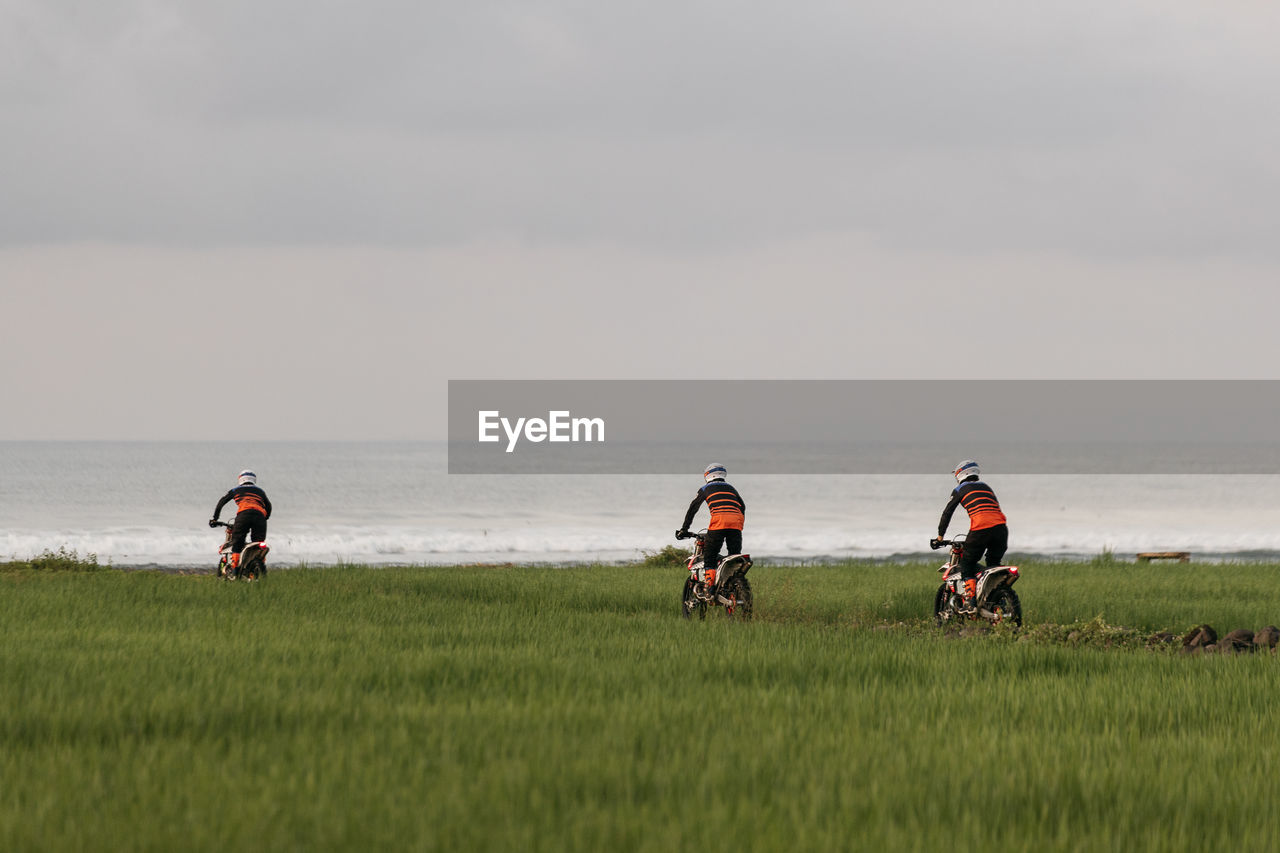 People riding dirt bikes on grass by sea against sky