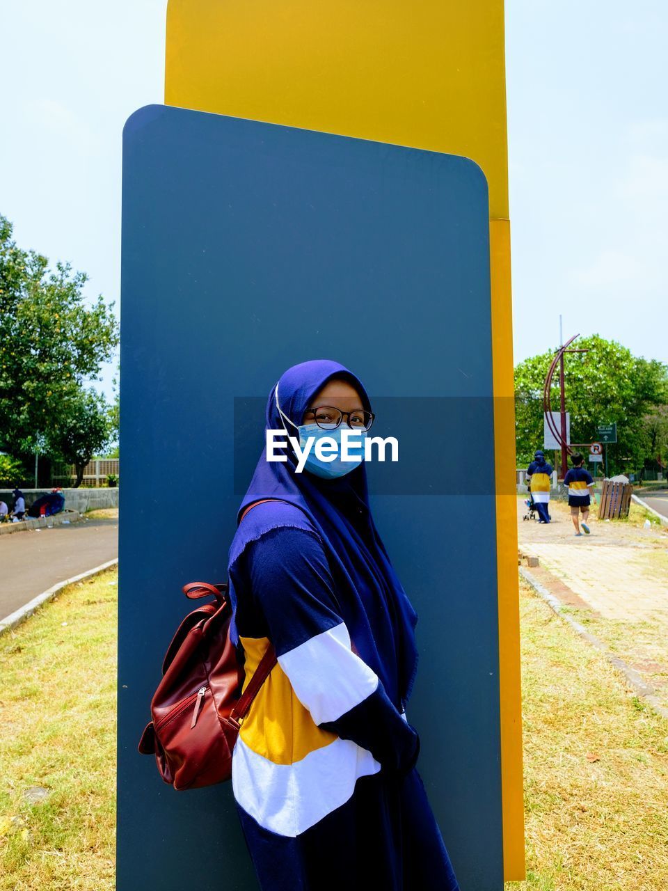 A girl wearing a blue hijab, a mask and a muslim casual tshirt standing on an empty blue billboard