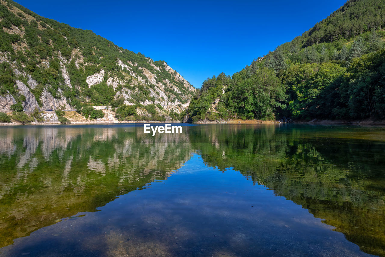 water, reflection, scenics - nature, lake, beauty in nature, mountain, tree, body of water, sky, plant, tranquility, nature, tranquil scene, environment, forest, landscape, blue, land, wilderness, reservoir, no people, pine tree, mountain range, pine woodland, coniferous tree, clear sky, pinaceae, non-urban scene, travel destinations, idyllic, travel, outdoors, day, woodland, tourism, green, summer, standing water, reflection lake, sunny