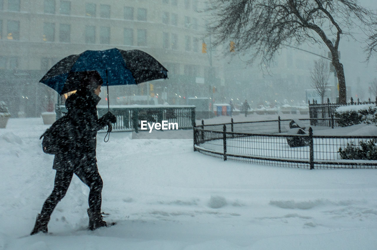 Side view of man with umbrella walking on snowy street during snowfall