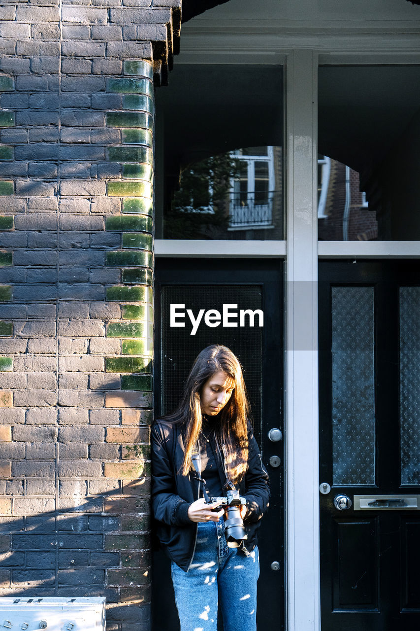 Concentrated young ethnic female traveler with long dark hair in casual outfit leaning on brick wall of aged building and watching photos on digital camera on sunny day in amsterdam