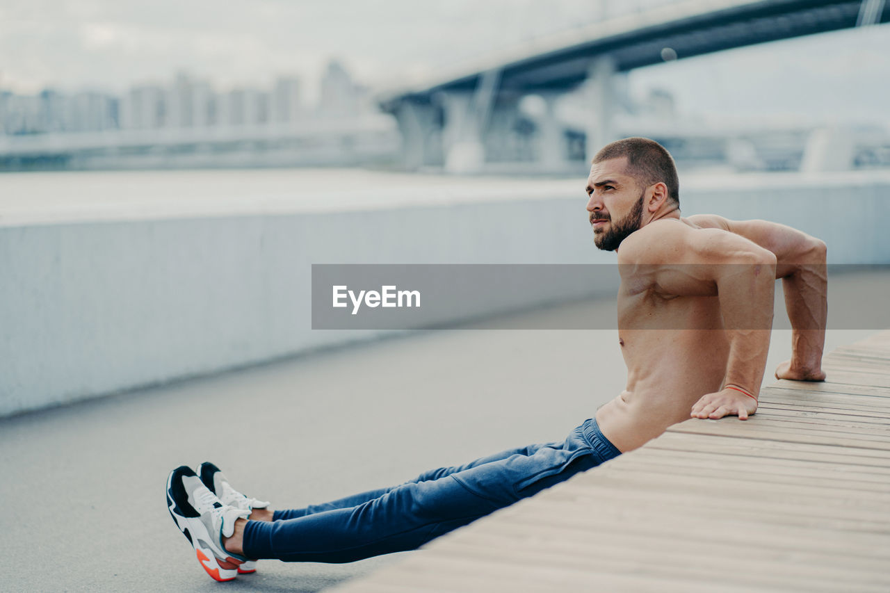 Side view of shirtless young man exercising on bridge