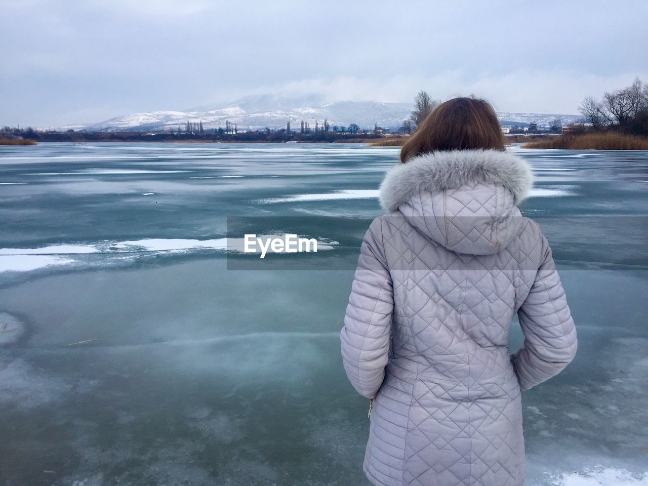 Rear view of woman standing at frozen lake against cloudy sky during winter
