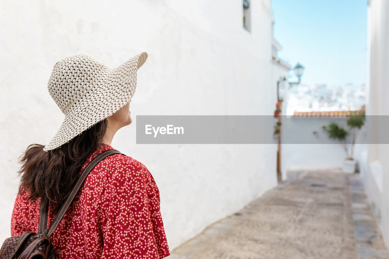 Side view of cheerful female traveler in straw hat on walkway between old buildings in white village in rhodes