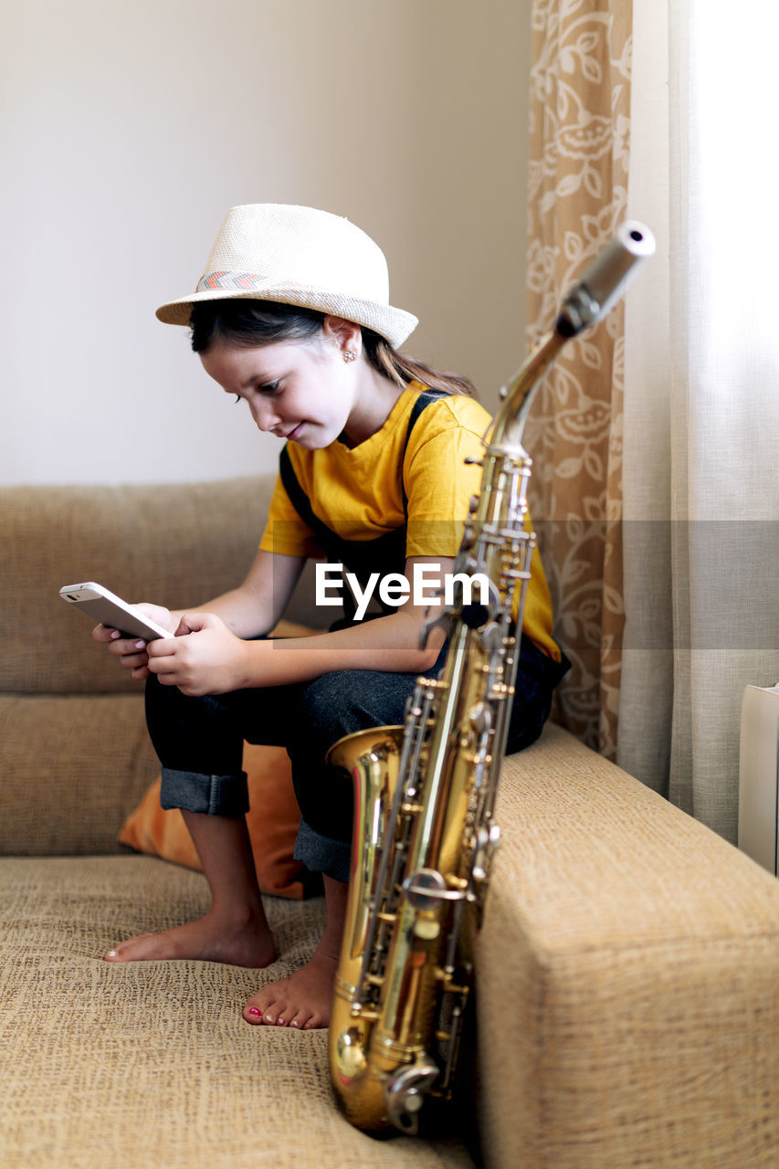 Child in hat text messaging on cellphone while sitting on couch with saxophone in living room