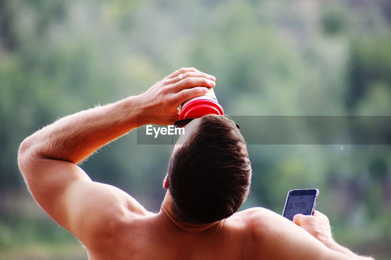 Rear view of shirtless man drinking while using mobile phone