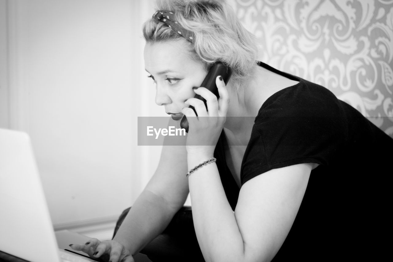 Woman talking on phone while using laptop at home