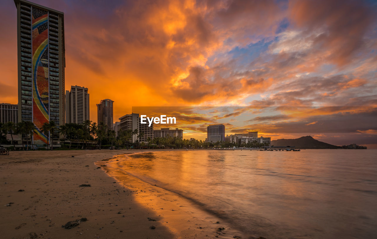 SCENIC VIEW OF BEACH BY BUILDINGS AGAINST SKY DURING SUNSET