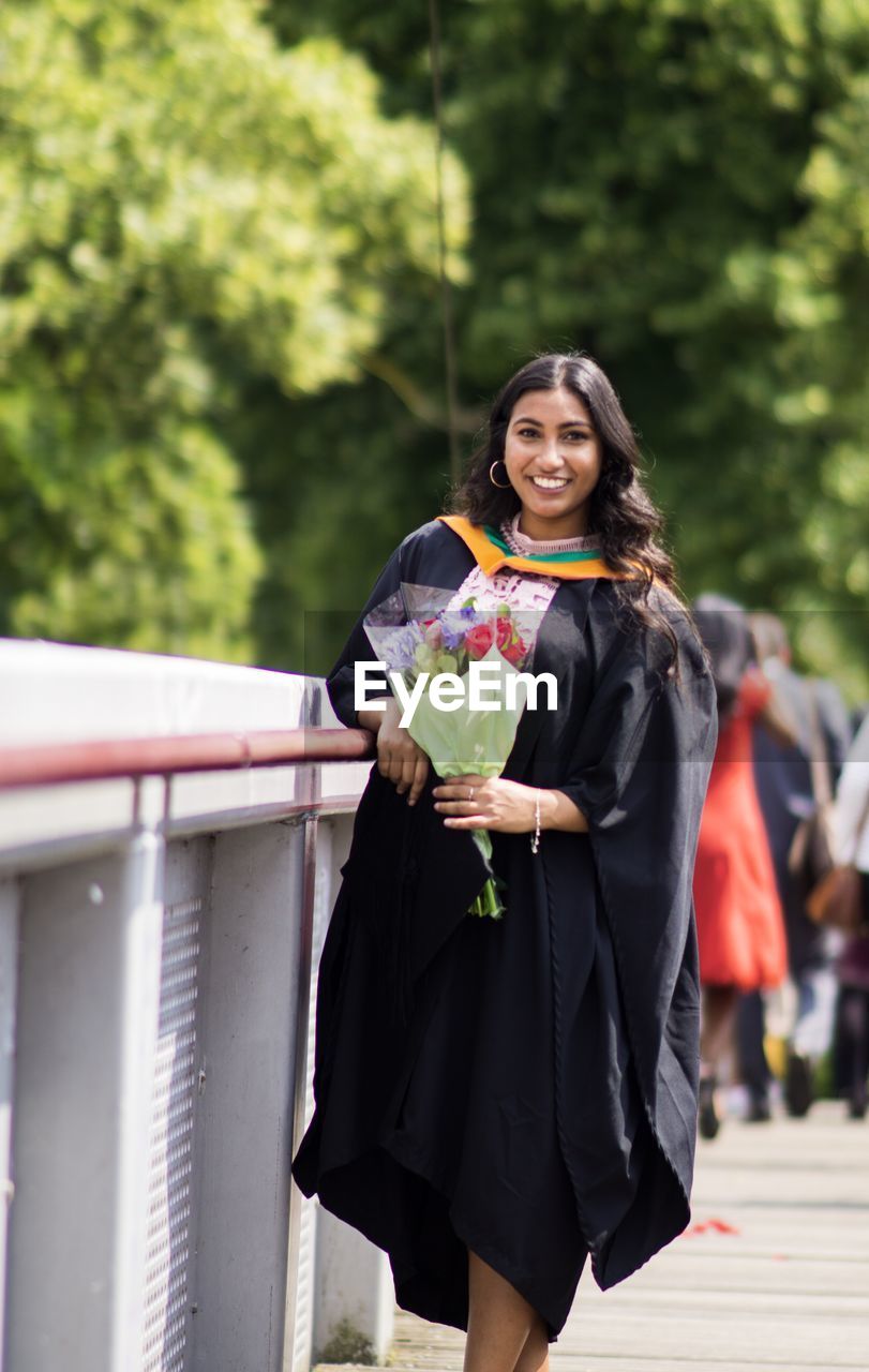 Portrait of young woman wearing graduation gown while standing on footpath against trees