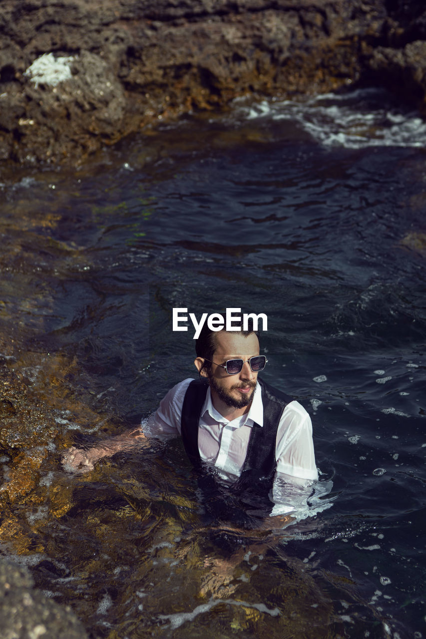 Man with a beard and sunglasses in clothes a vest and a white shirt swims in the sea among the rocks