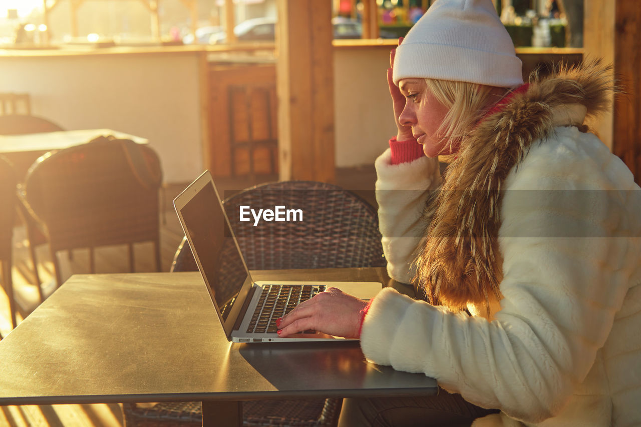 A woman in a sweater and a fur coat is sitting on the terrace of a cafe with a laptop.