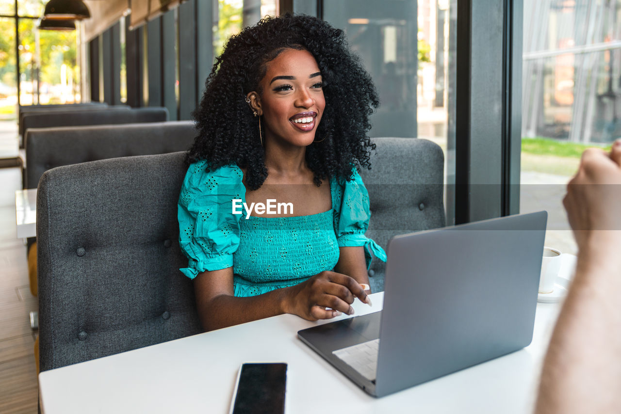 Cheerful young african american woman in turquoise top sitting at table with smartphone and working via netbook while chatting with cropped unrecognizable boyfriend in drinking coffee