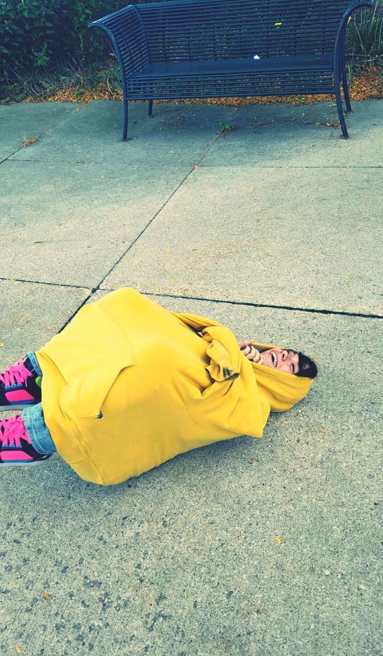 High angle view of playful woman covered with yellow hooded shirt lying on footpath