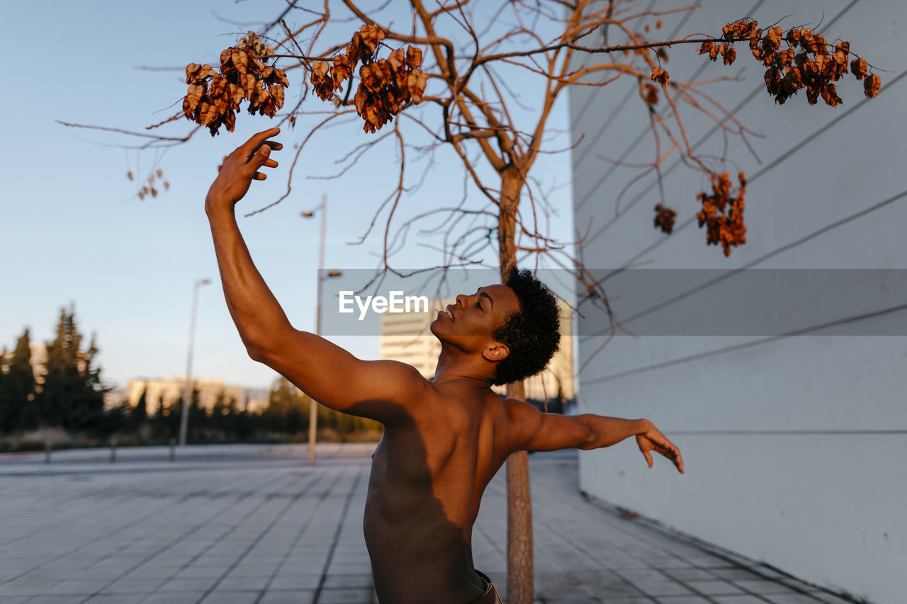 Shirtless man dancing against bare tree in city