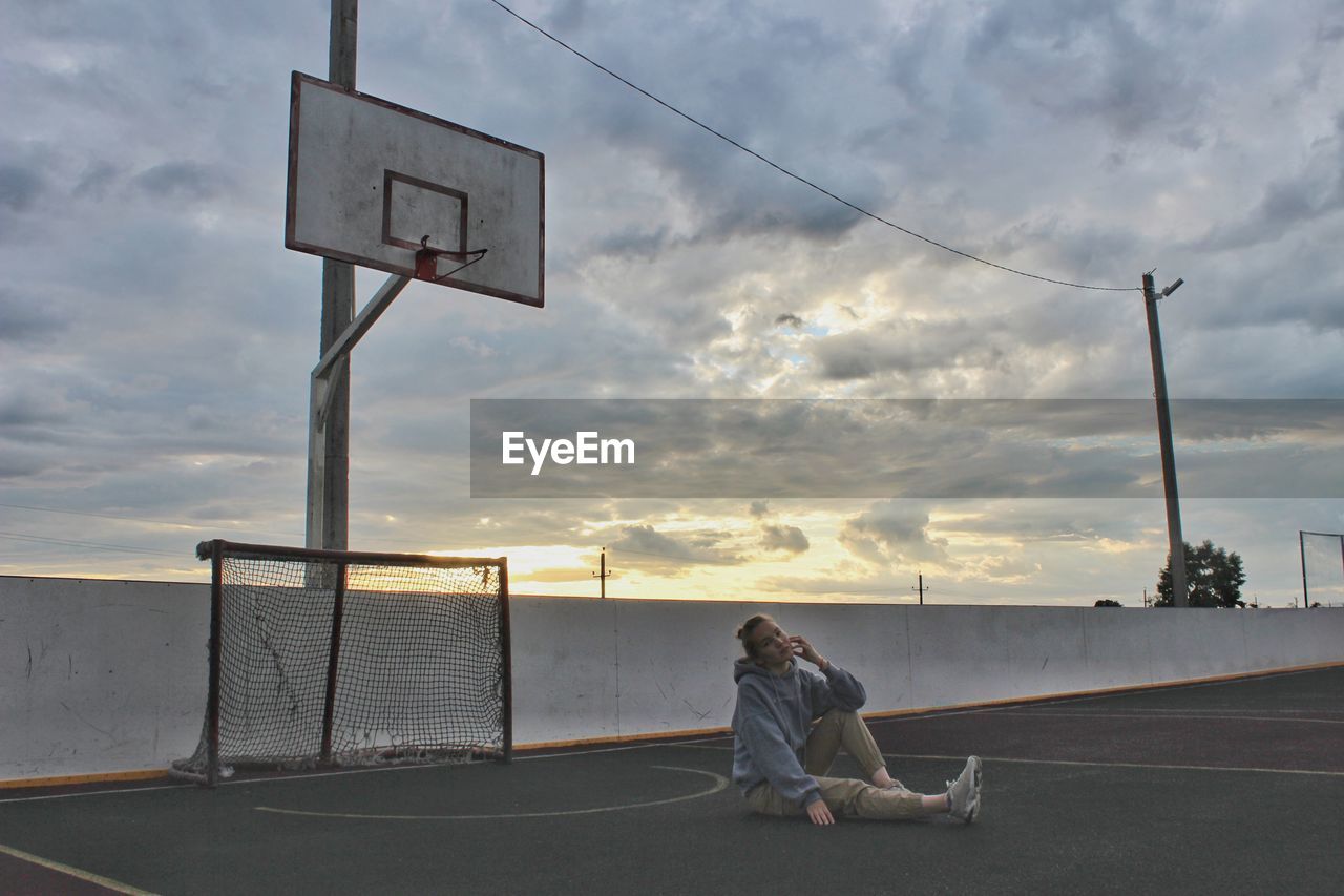 Woman sitting in sports court against cloudy sky during sunset