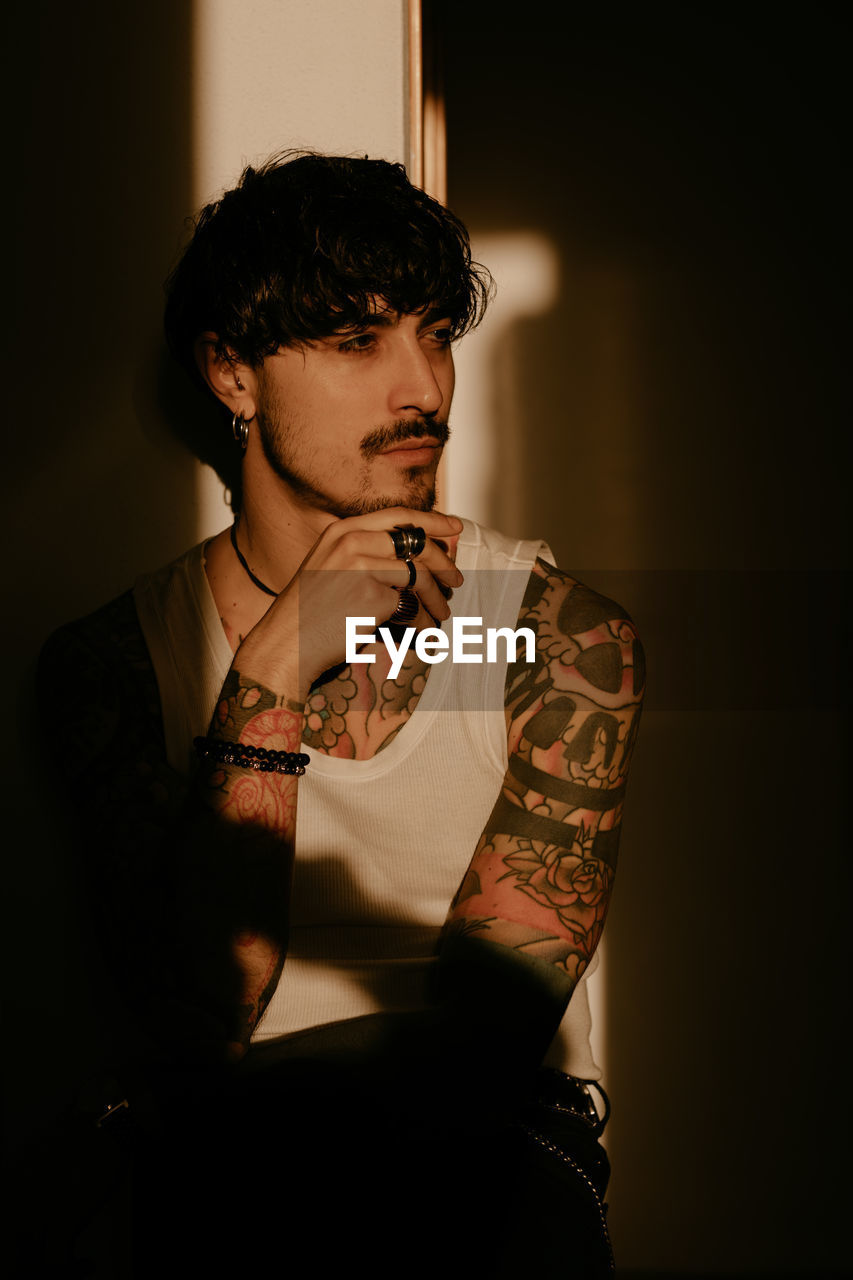 Young stylish guy with mustache and tattoos wearing white tank top and leaning against wall with back while looking away