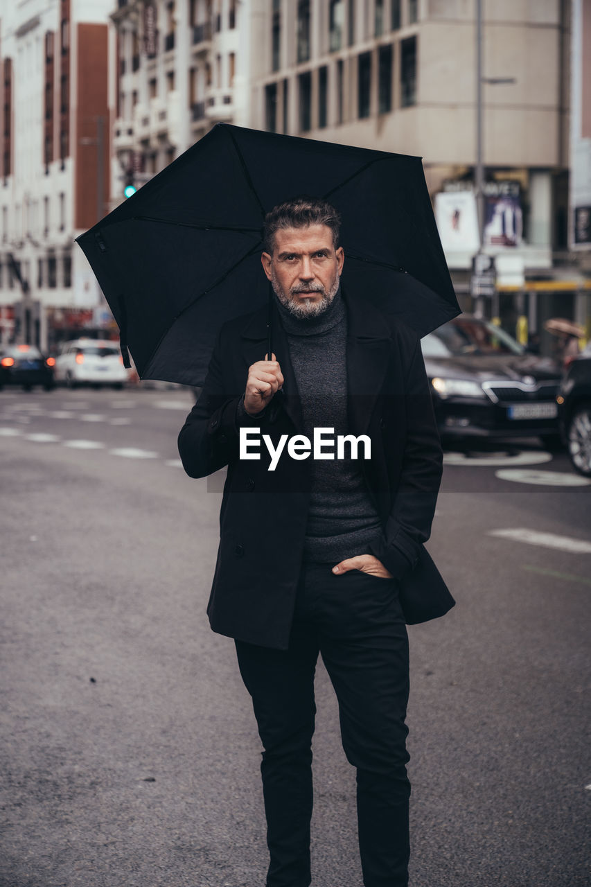 Pensive successful mature bearded male entrepreneur in black standing under umbrella during walk in street in rainy day