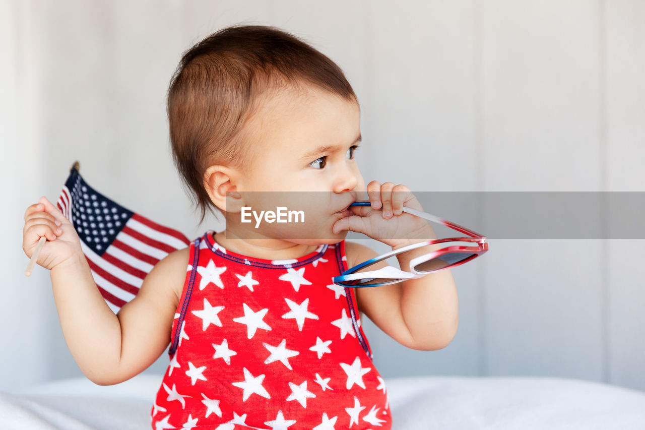 Cute baby girl holding sunglasses and american flag while sitting on bed at home