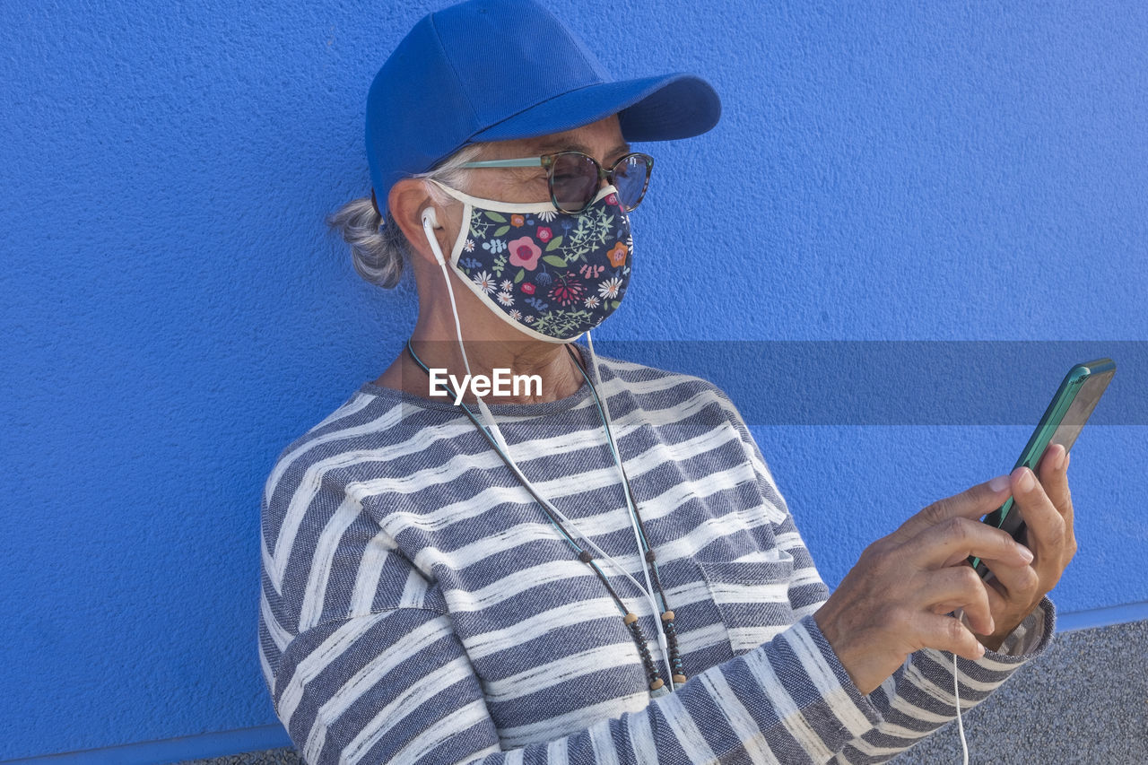 Close-up of senior woman wearing sunglasses using smart phone standing against blue wall